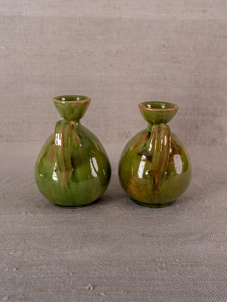 Pair of antique French green glazed earthenware pitchers with narrow openings on beige textured surface.