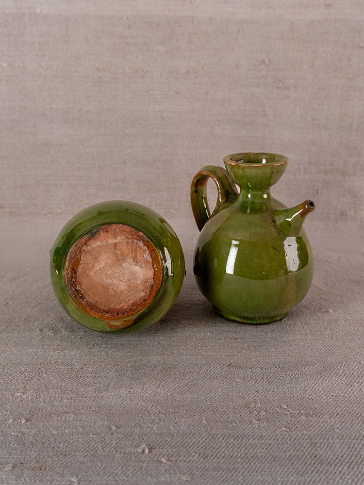 Pair of antique French green-glazed earthenware pitchers on textured grey fabric, one upright, one on its side.