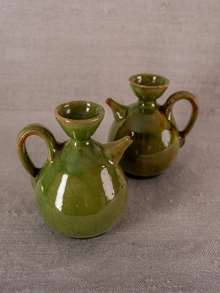 Pair of antique French green glazed earthenware pitchers with handles and spouts on gray textured surface.