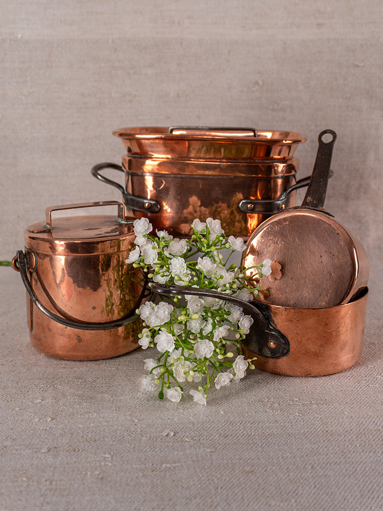 19th-century French copper apprentice daubiere and saucepans arranged with white flowers on neutral fabric.