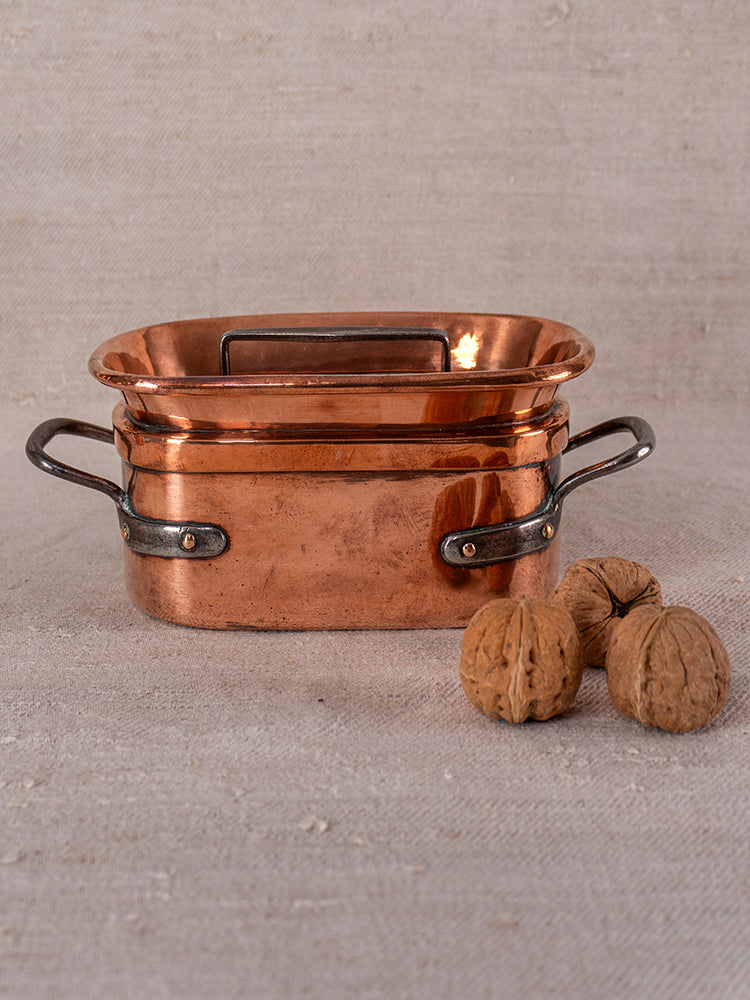 19th-century French copper daubiere and saucepans with dark handles on textured beige; three walnuts at right.