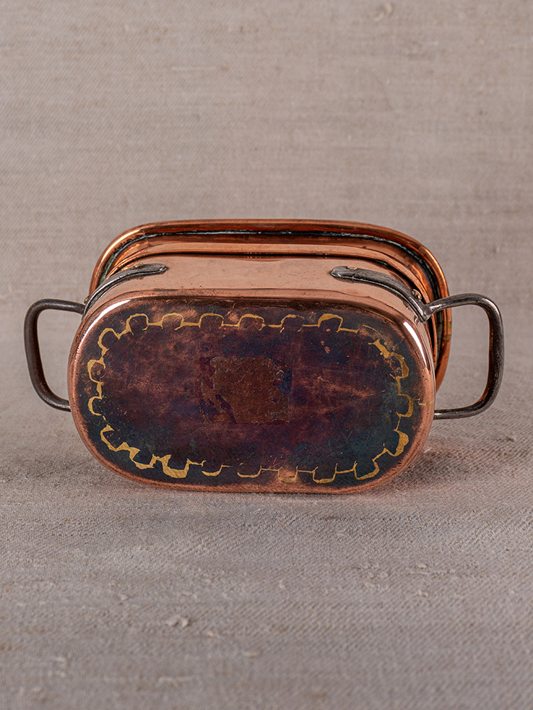 Antique French copper apprentice daubiere with dark metal handles on textured neutral fabric background.