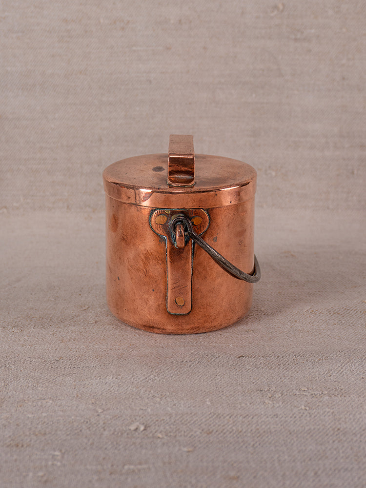 Antique French copper daubiere and saucepans with lid and metal handle on textured beige fabric, showing graceful tarnish.