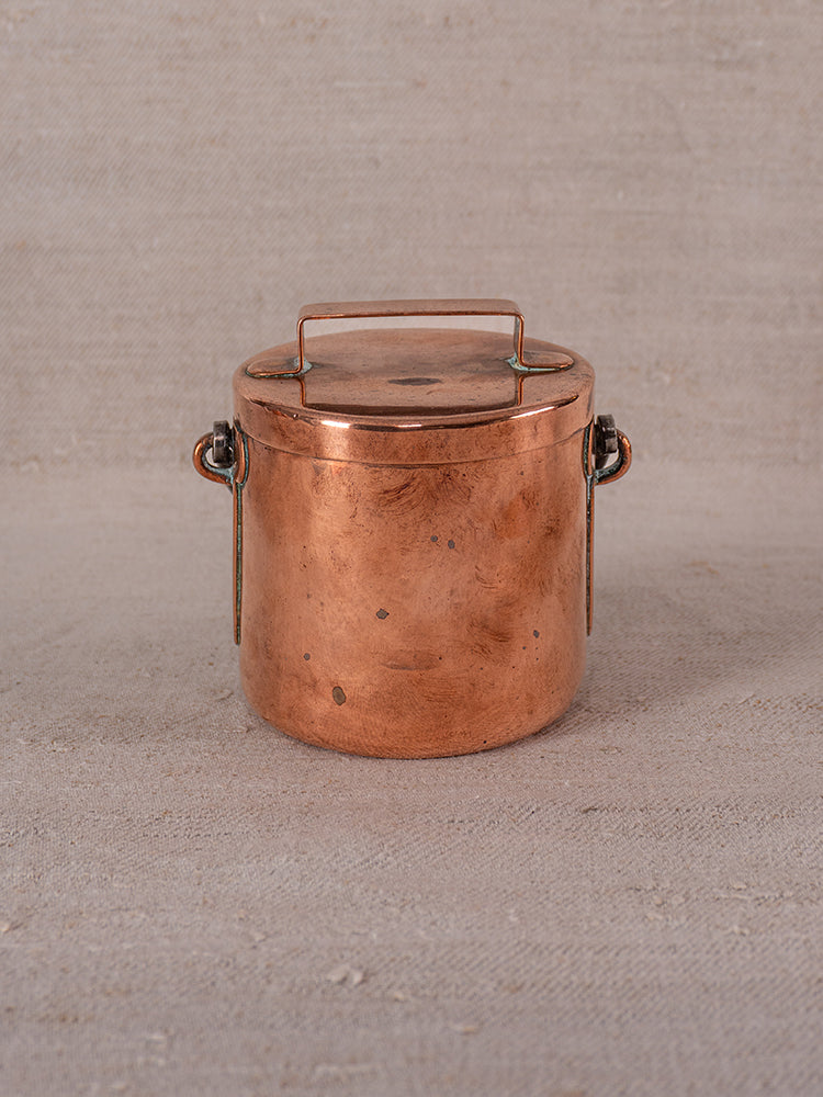 Antique French copper apprentice daubiere and saucepans with handled lid and side clasps on beige fabric surface.