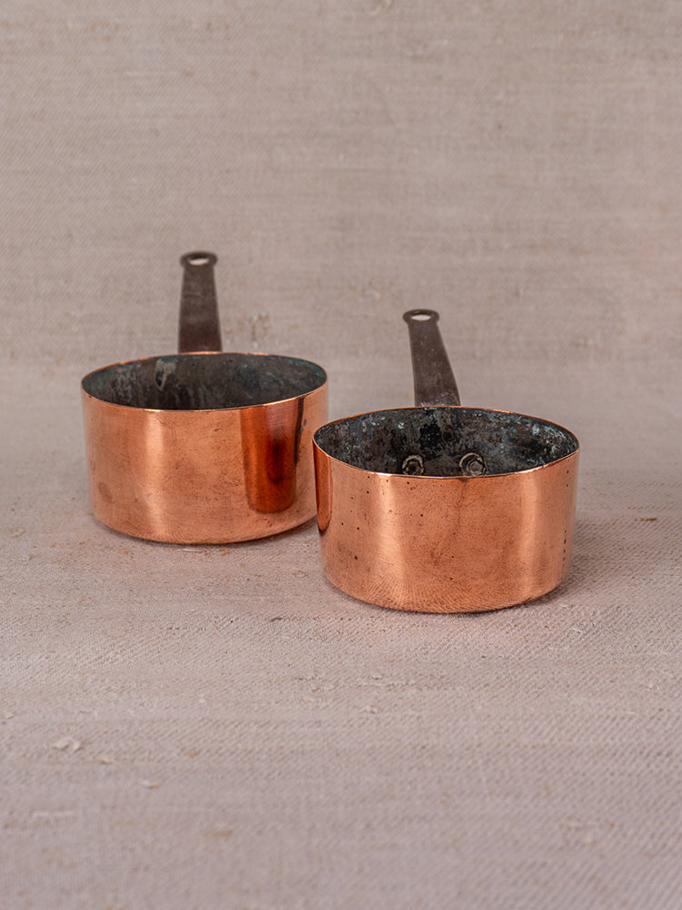 Antique French copper daubiere and saucepan set with dark interiors, shown on a textured beige surface.