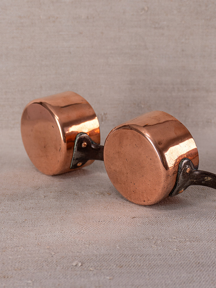 Pair of small round French copper saucepans with metal handles, polished finish, upside down on textured fabric.