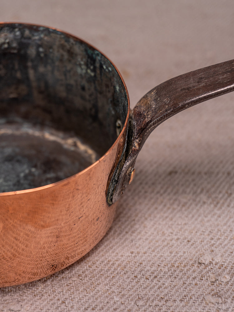Antique French copper saucepan with worn handle and tarnish, displayed on textured beige fabric.