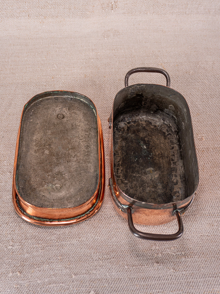 Antique French copper daubiere and saucepans with lid and side handles, aged patina, on textured beige fabric.
