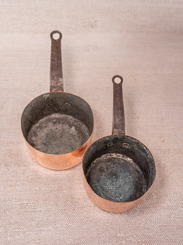 Pair of small antique French copper saucepans with worn interiors and hanging handles on textured fabric.