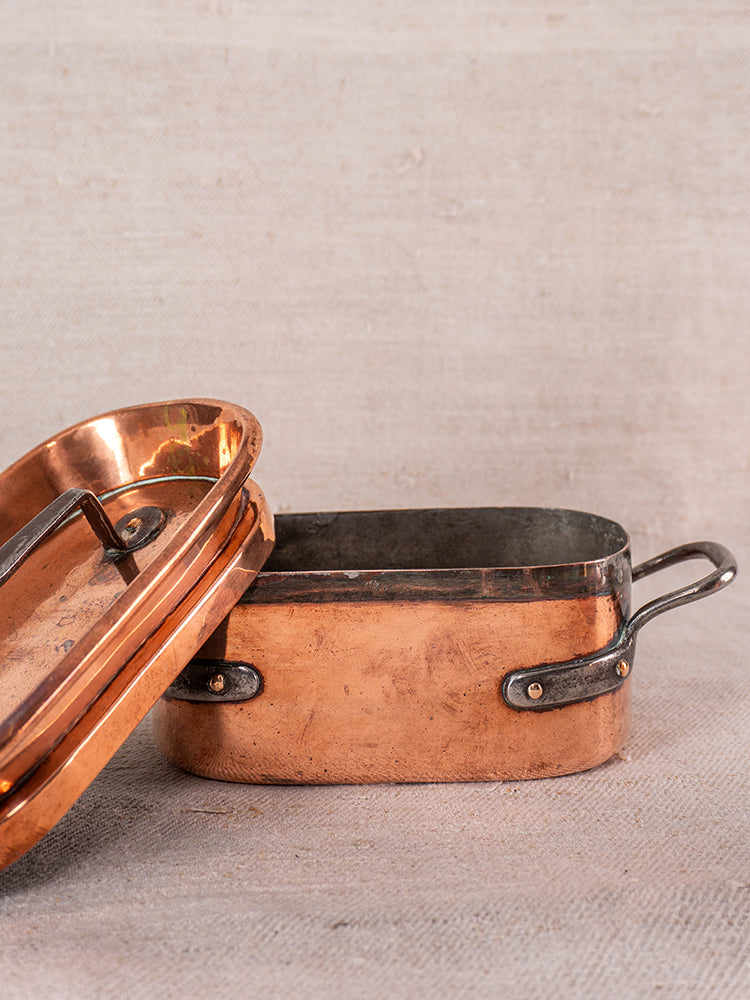 Antique French copper daubiere and saucepans with black handles and lid on a textured neutral surface.