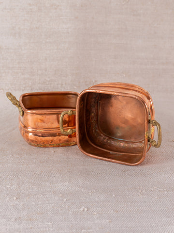 Pair of antique French hammered copper pots with brass handles, one upright and one on their side, on light beige textured fabric.