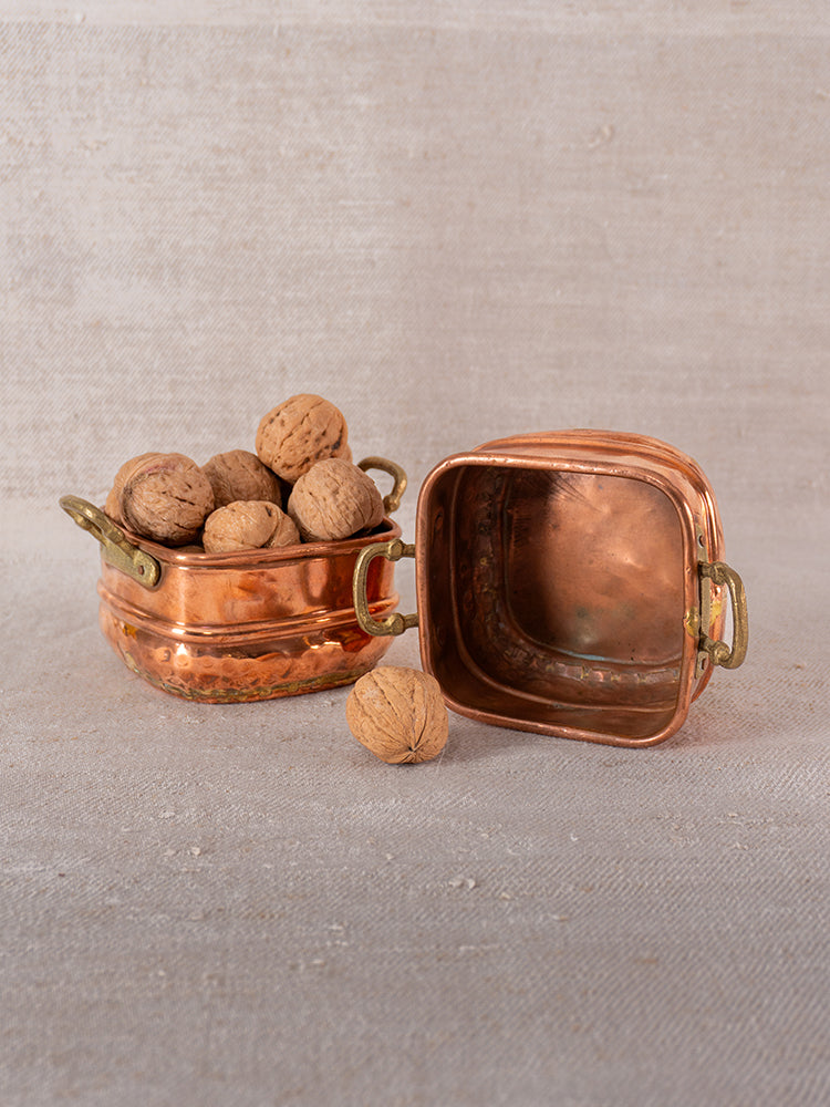 Antique French hammered copper pot with brass handles, filled with walnuts, lid and walnut on beige textured surface.