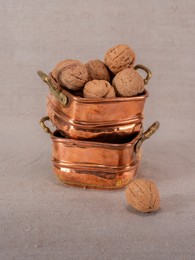 Antique French hammered copper pots with brass handles, stacked on beige textured surface, top pot filled with walnuts.