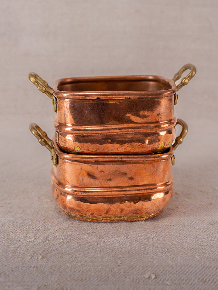 Antique French hammered copper pots with brass handles stacked on neutral fabric background, late 19th to early 20th century.