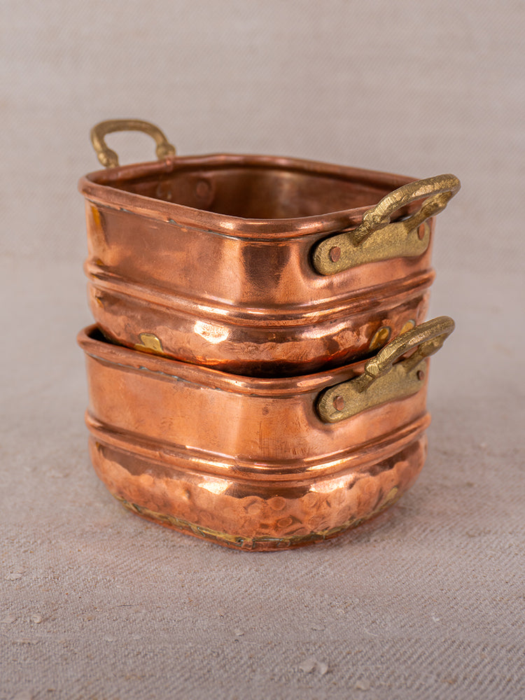 Pair of antique French hammered copper pots with brass handles, stacked on a neutral fabric background.