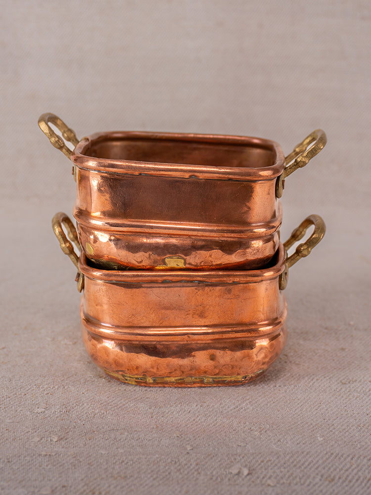 Pair of antique French hammered copper pots with brass handles and dovetail seams stacked on neutral fabric.