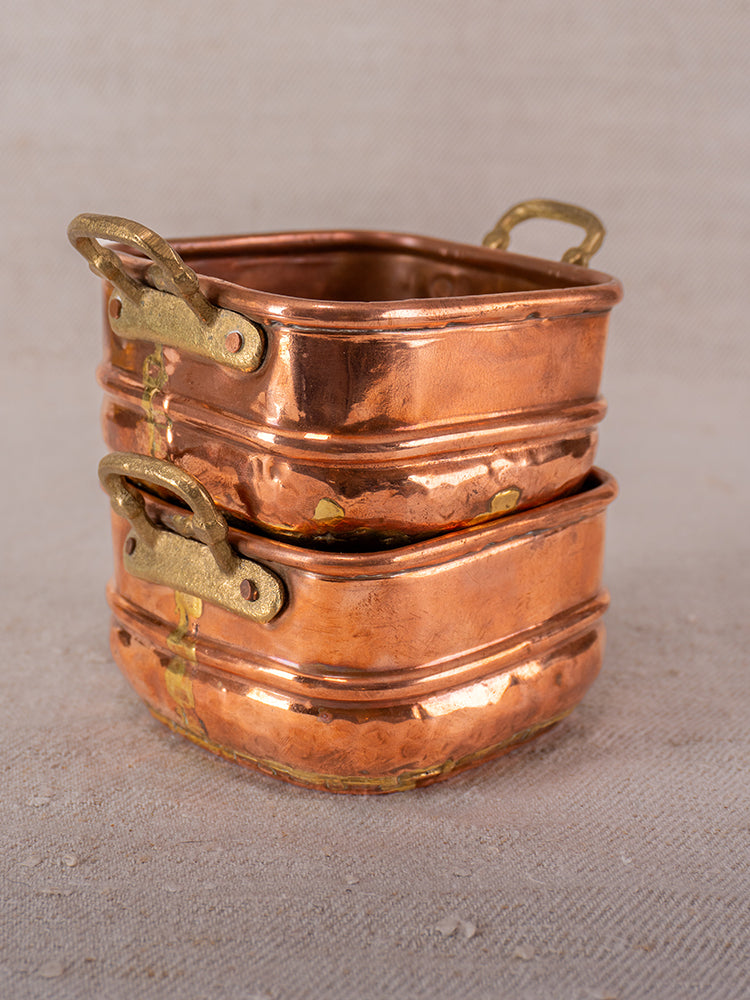 Antique French copper pots with hammered finish, brass handles, and dovetail seams stacked on a neutral textured backdrop.