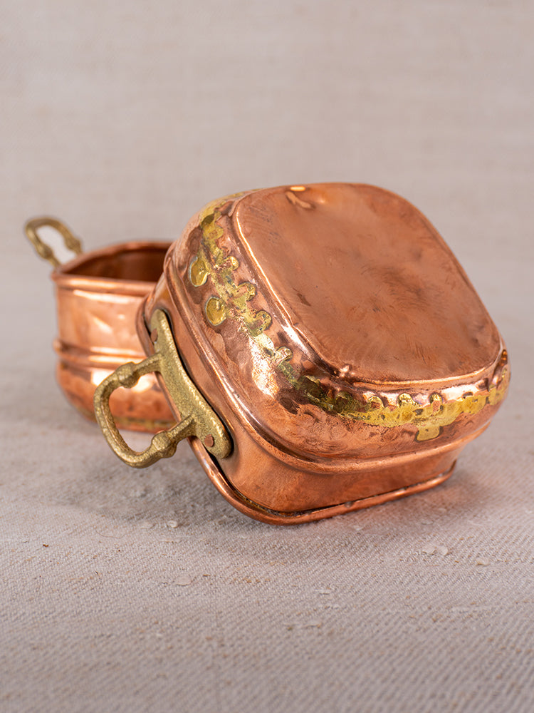 Antique French hammered copper pots with brass handles, one open and one closed, showcasing brass dovetail seams on fabric.