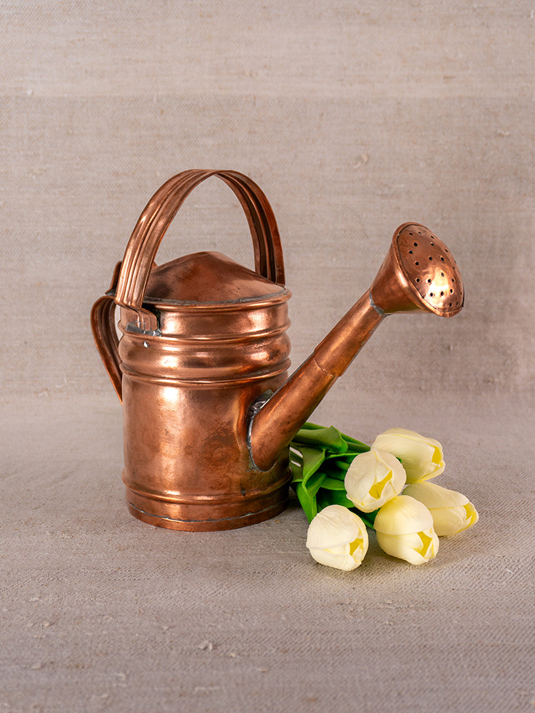 Antique French copper watering can with riveted handle, displayed on beige fabric next to fresh white tulips.