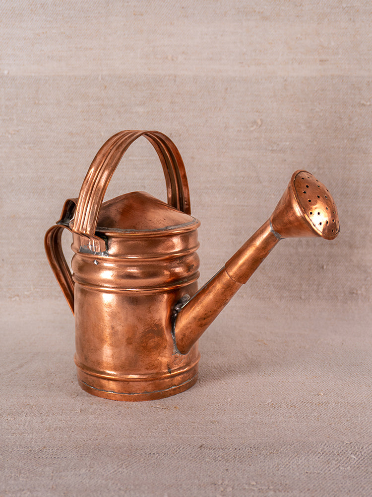 Antique French copper watering can with curved handle and long spout on a neutral fabric background.