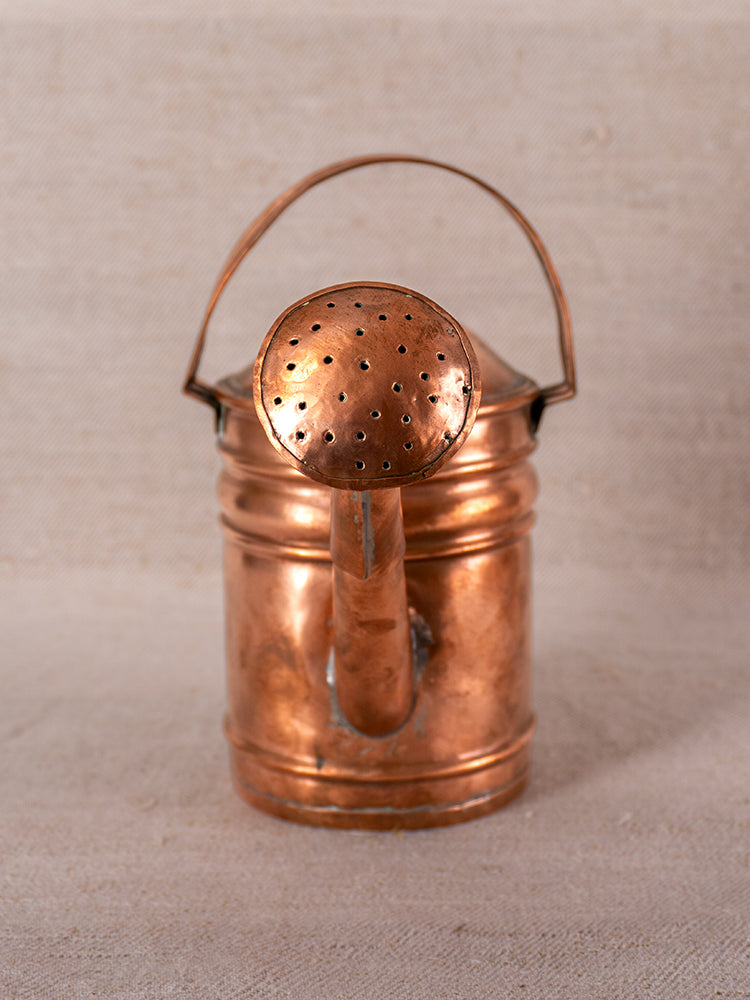 Antique French copper watering can with riveted design, rounded handle, and perforated spout on neutral fabric background.