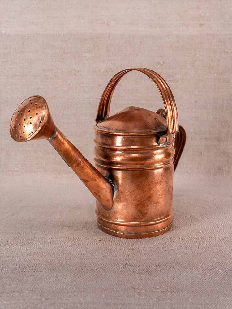 Antique French copper watering can with long spout and rounded handle on a neutral surface, beige background.