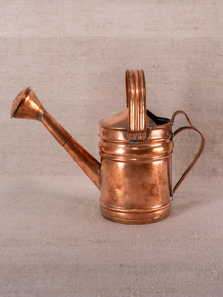 Antique French copper watering can with riveted details and long spout on textured beige fabric.