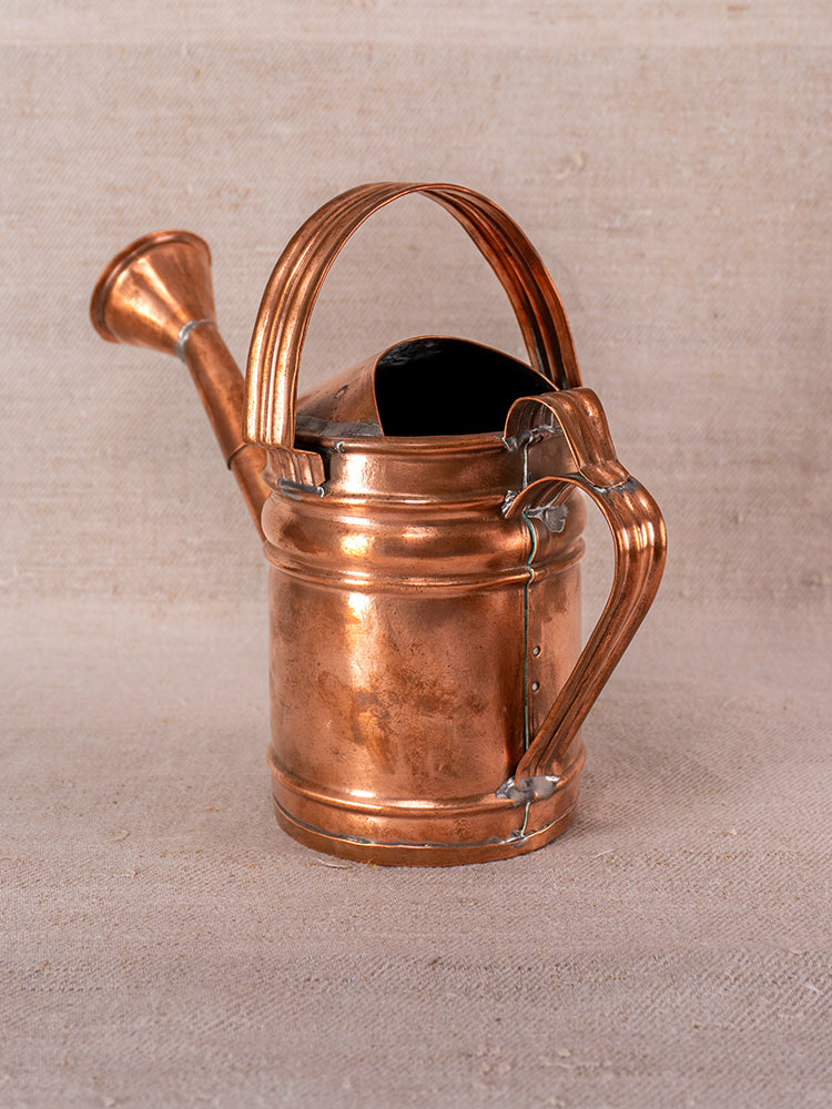 Antique French copper watering can with riveted details, rounded handle, and curved spout on neutral fabric surface.