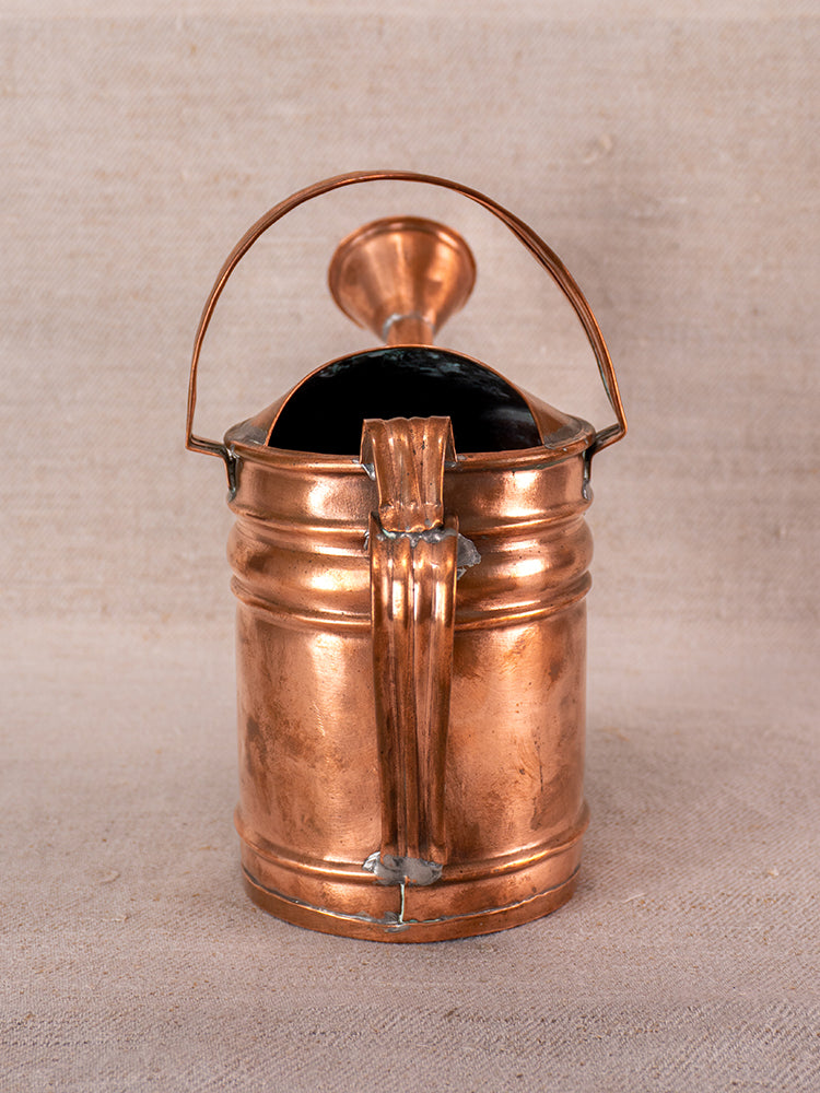 Antique French copper watering can with curved handle, spout, and ridged details on neutral fabric background.