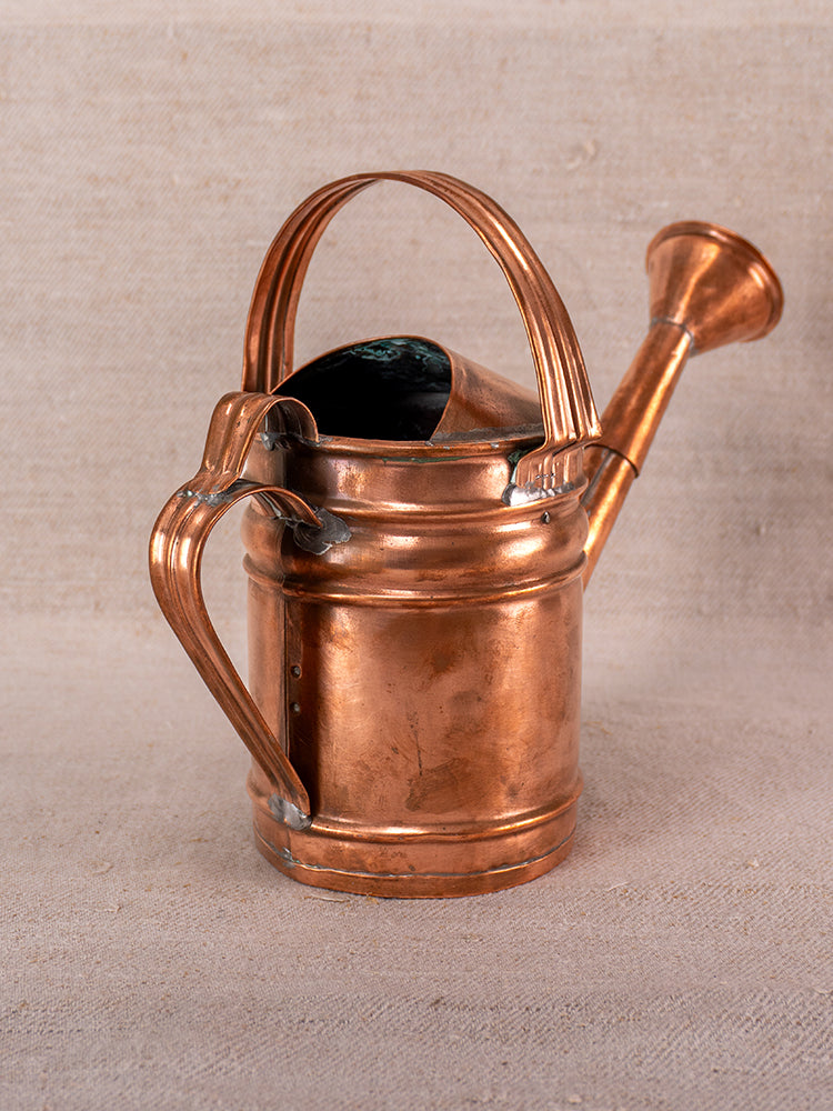 Antique French copper watering can with large handle and spout on textured beige fabric.