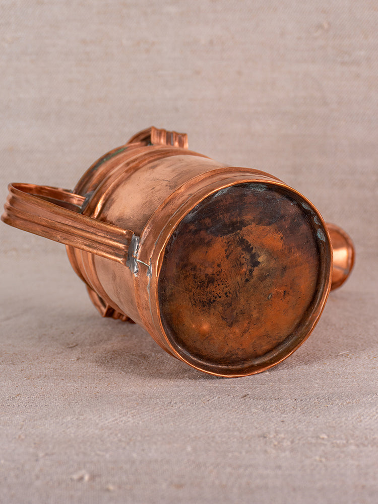 Antique French copper watering can with visible patina displayed on textured beige fabric backdrop.
