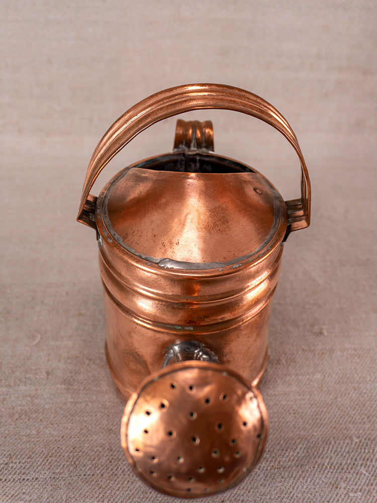 Antique French copper watering can with curved handle and large spout on neutral textured surface.