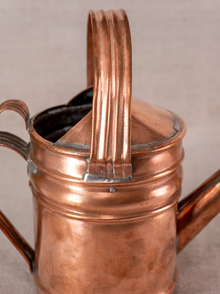 Antique French copper watering can with curved handle, spout, and rich patina on textured fabric background.