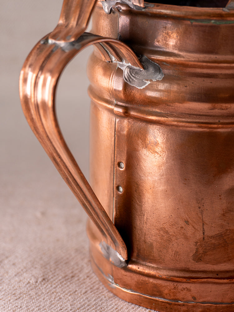 Antique French copper watering can with riveted seams, curved handle, warm patina, on neutral fabric background.