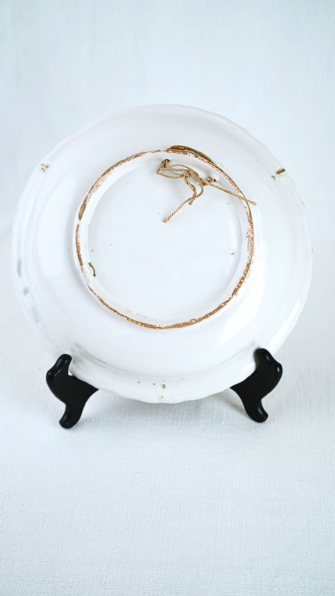 Underside of a French Faience Chinoiserie bowl, glazed white, resting on a black wire stand.