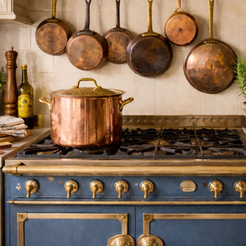 copper and metalware collection featuring antique French copper cookware displayed on a blue range with hanging pans and pot lid in a light-filled kitchen