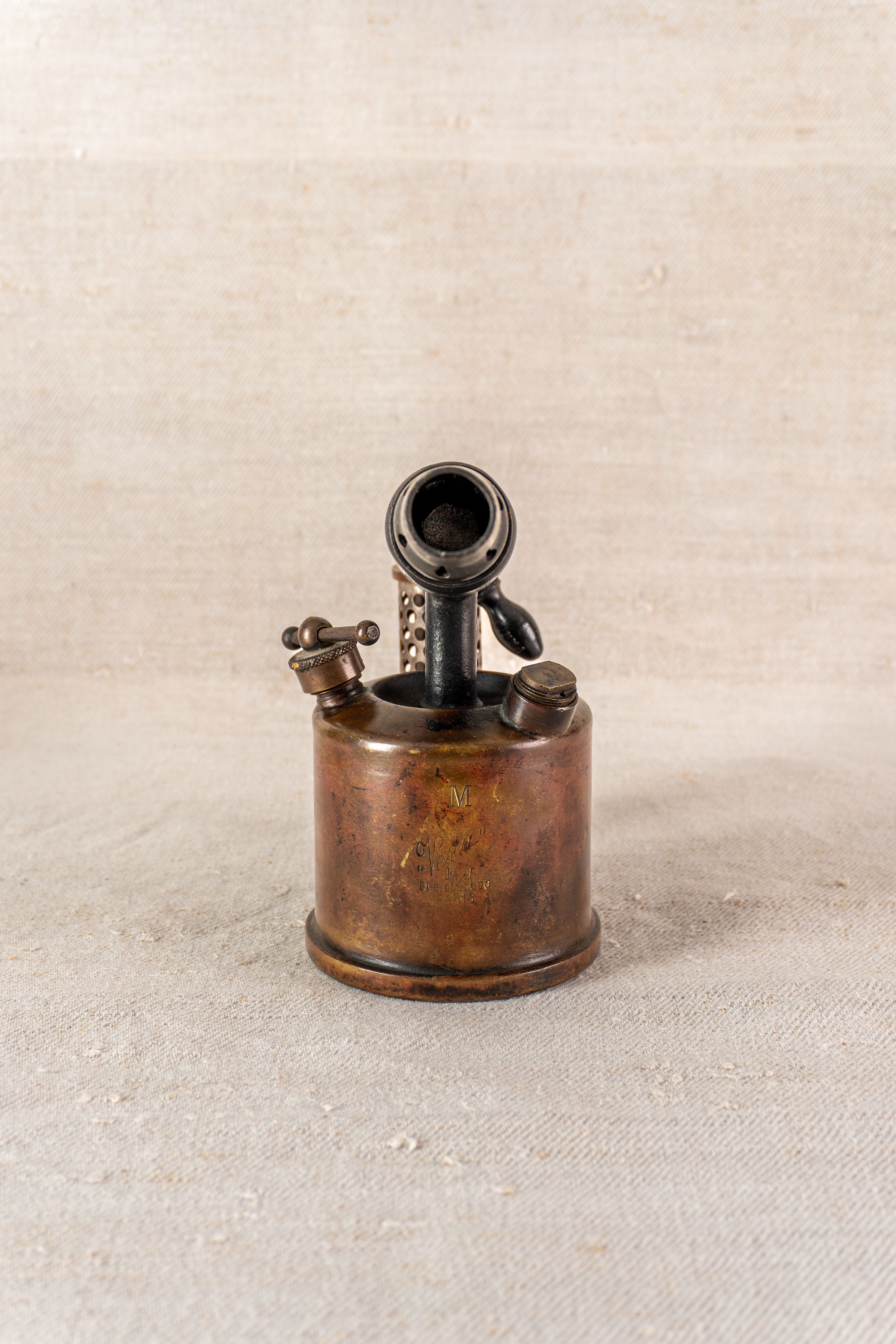 Vintage French blowtorch by Vesta F.J. Paris circa 1920, round fuel tank, upright on beige textured surface.