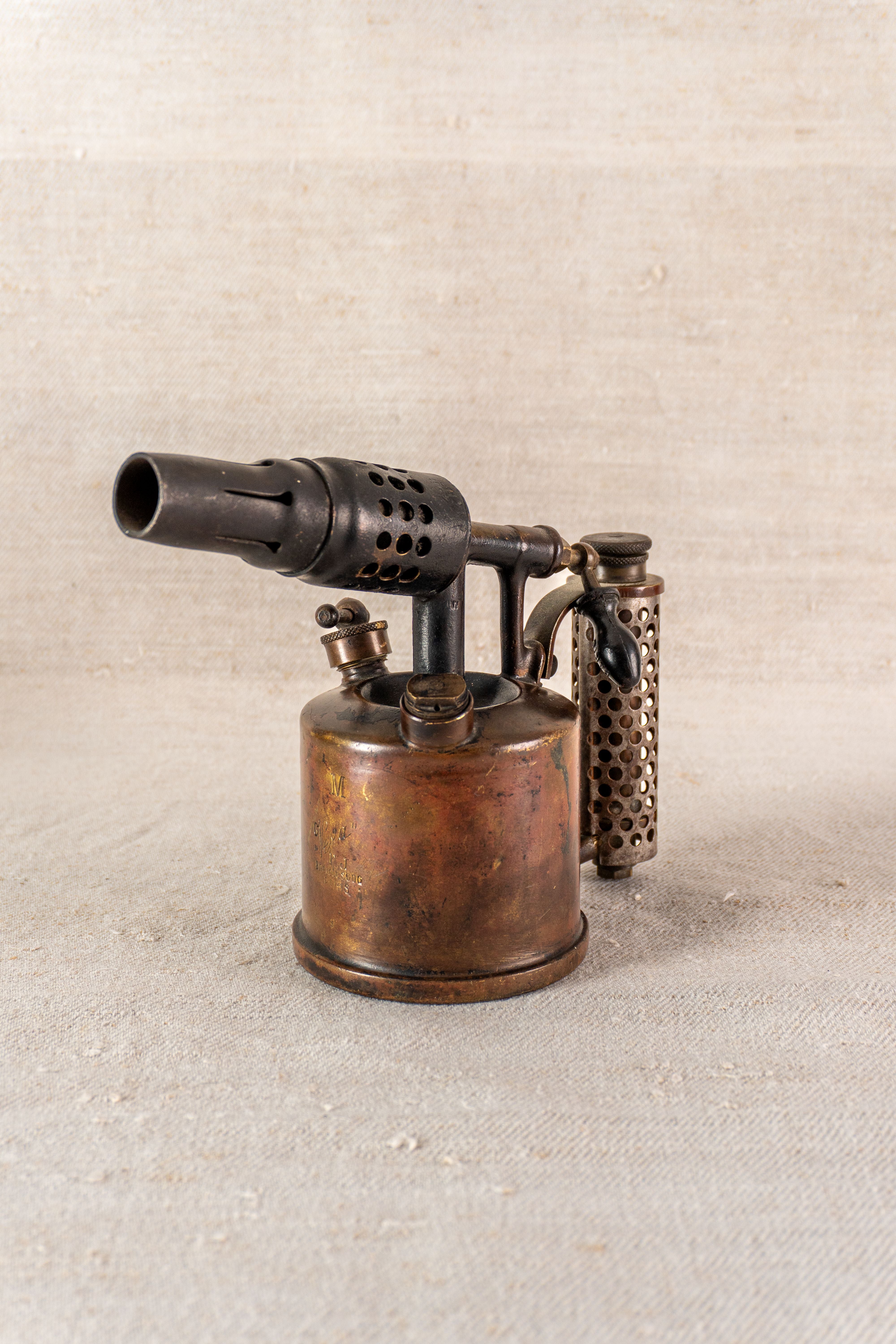 Vintage French blowtorch by Vesta F.J. Paris, circa 1920, brass body, perforated handle, neutral textured background.