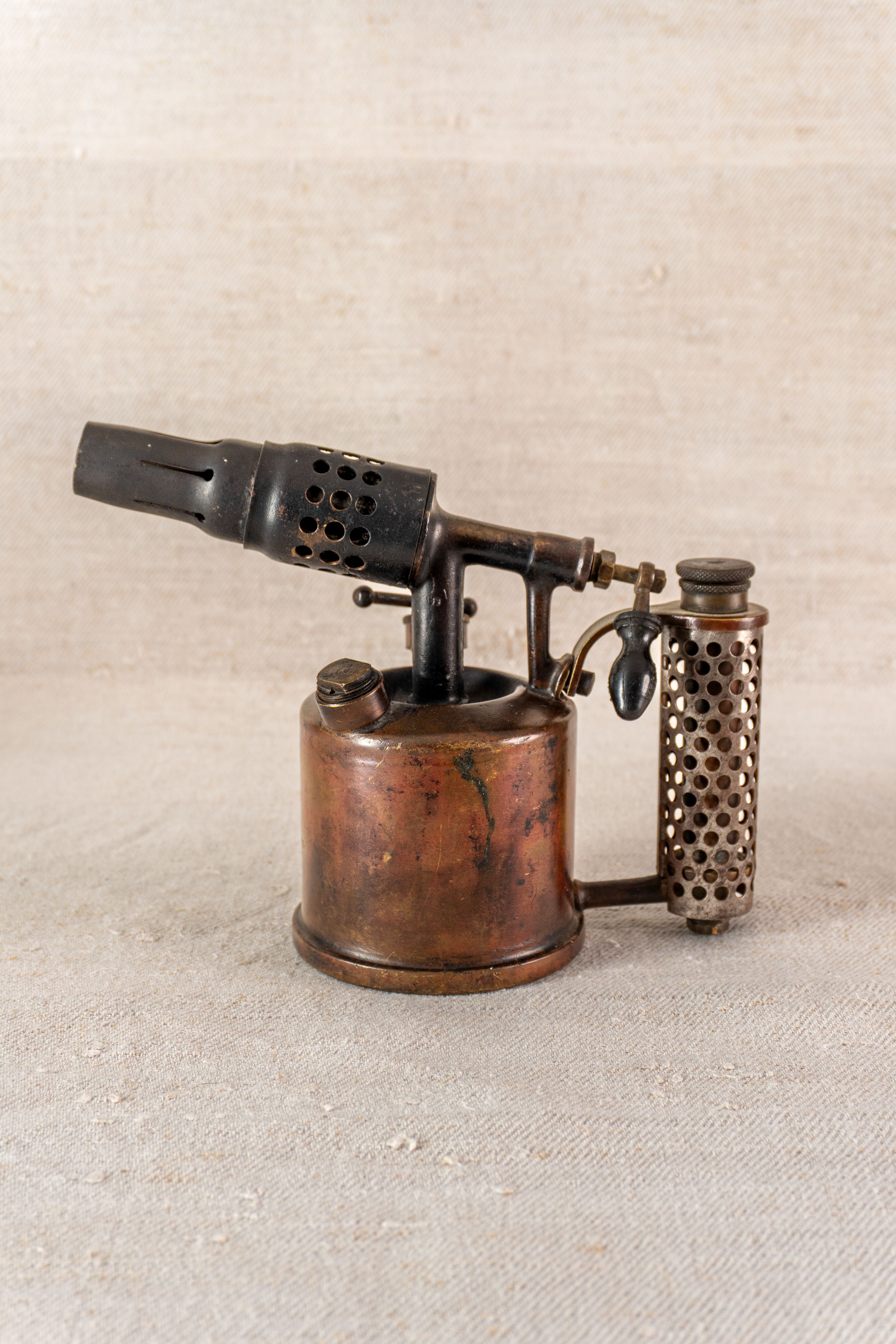 Vintage French Vesta F.J. Paris blowtorch, circa 1920, with cylindrical fuel tank and perforated handle on textured beige surface.