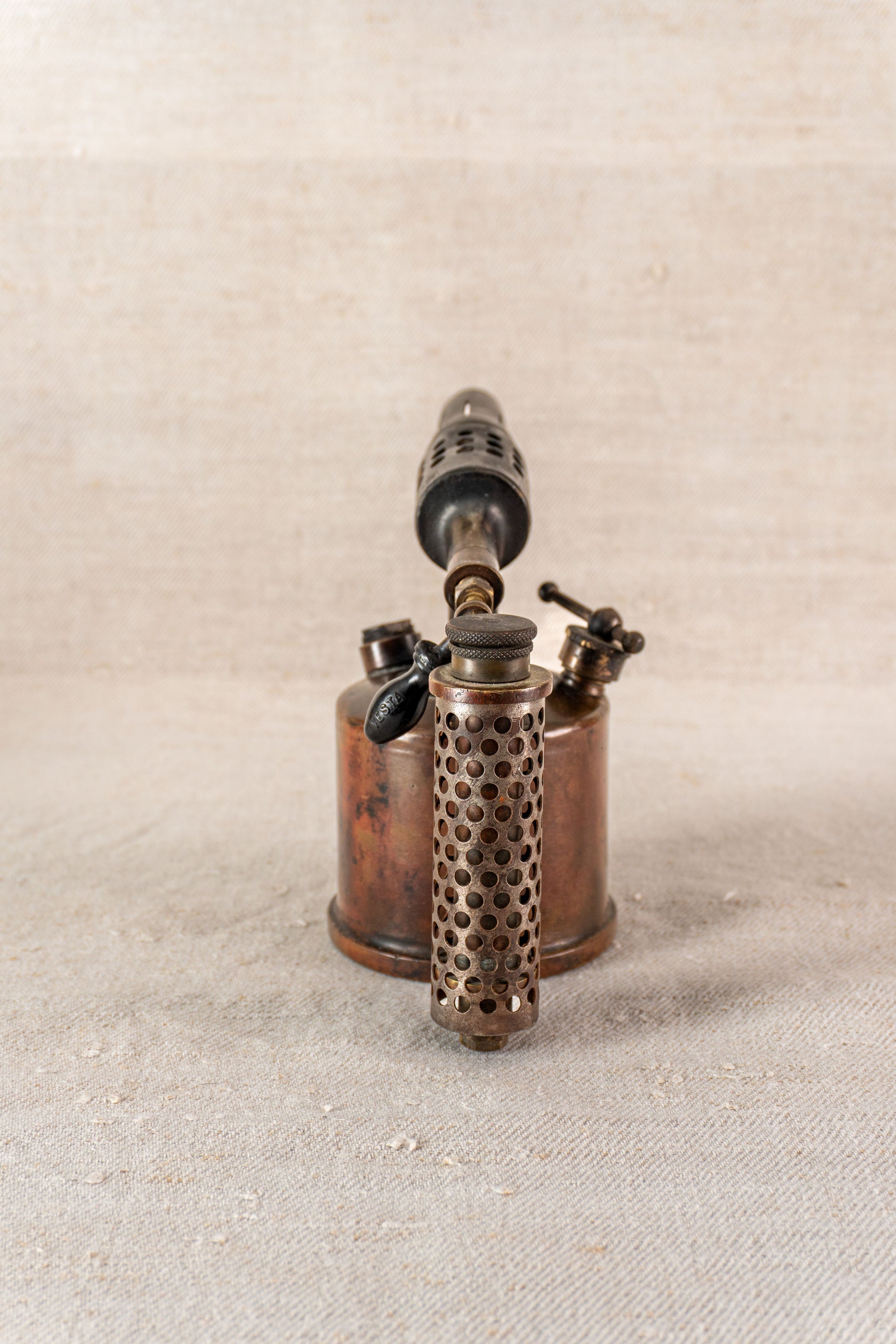 Vintage French Vesta F.J. Paris brass blowtorch, perforated cylindrical handle, upright on textured neutral surface.