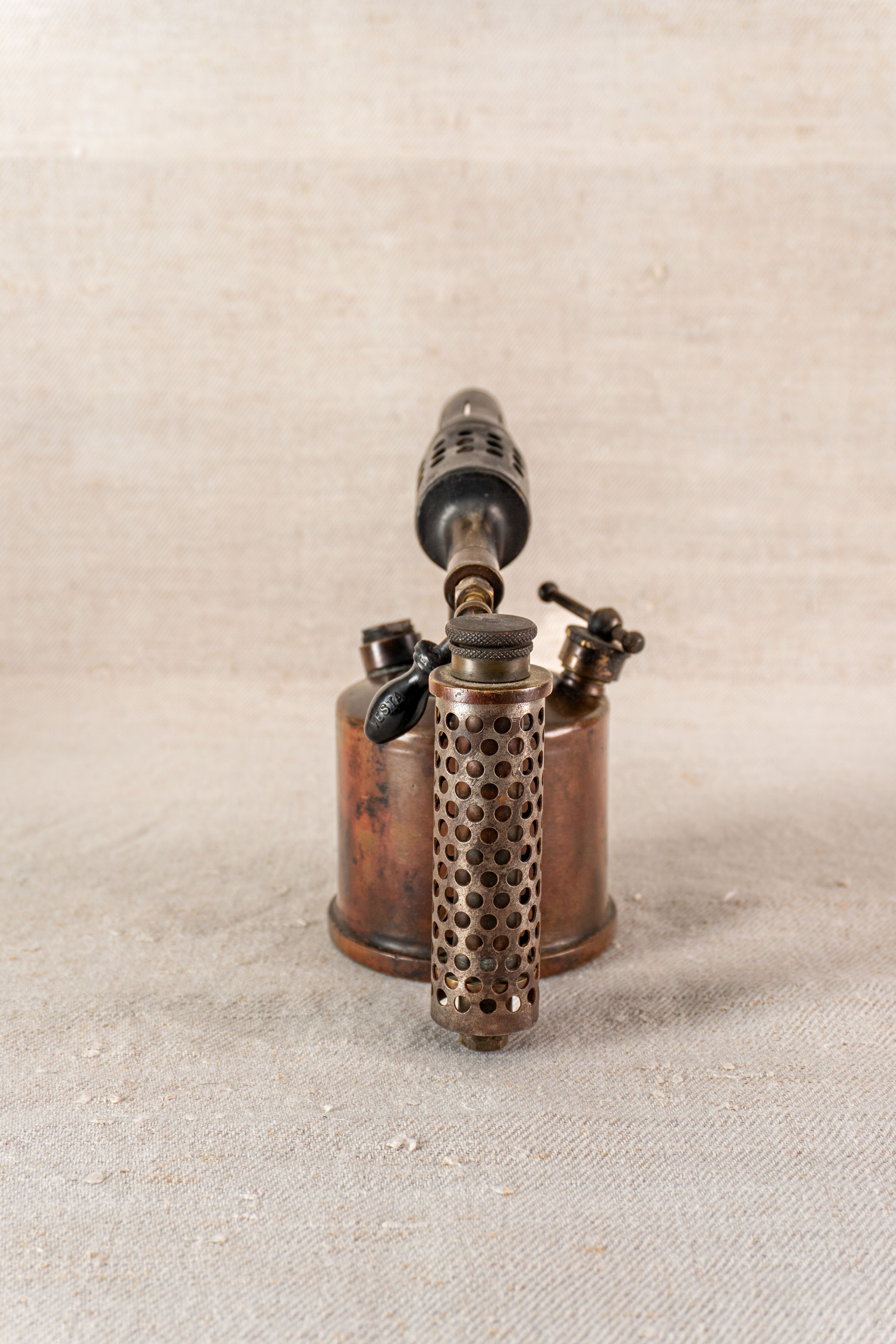 Vintage French Vesta F.J. Paris brass blowtorch, perforated cylindrical handle, upright on textured neutral surface.