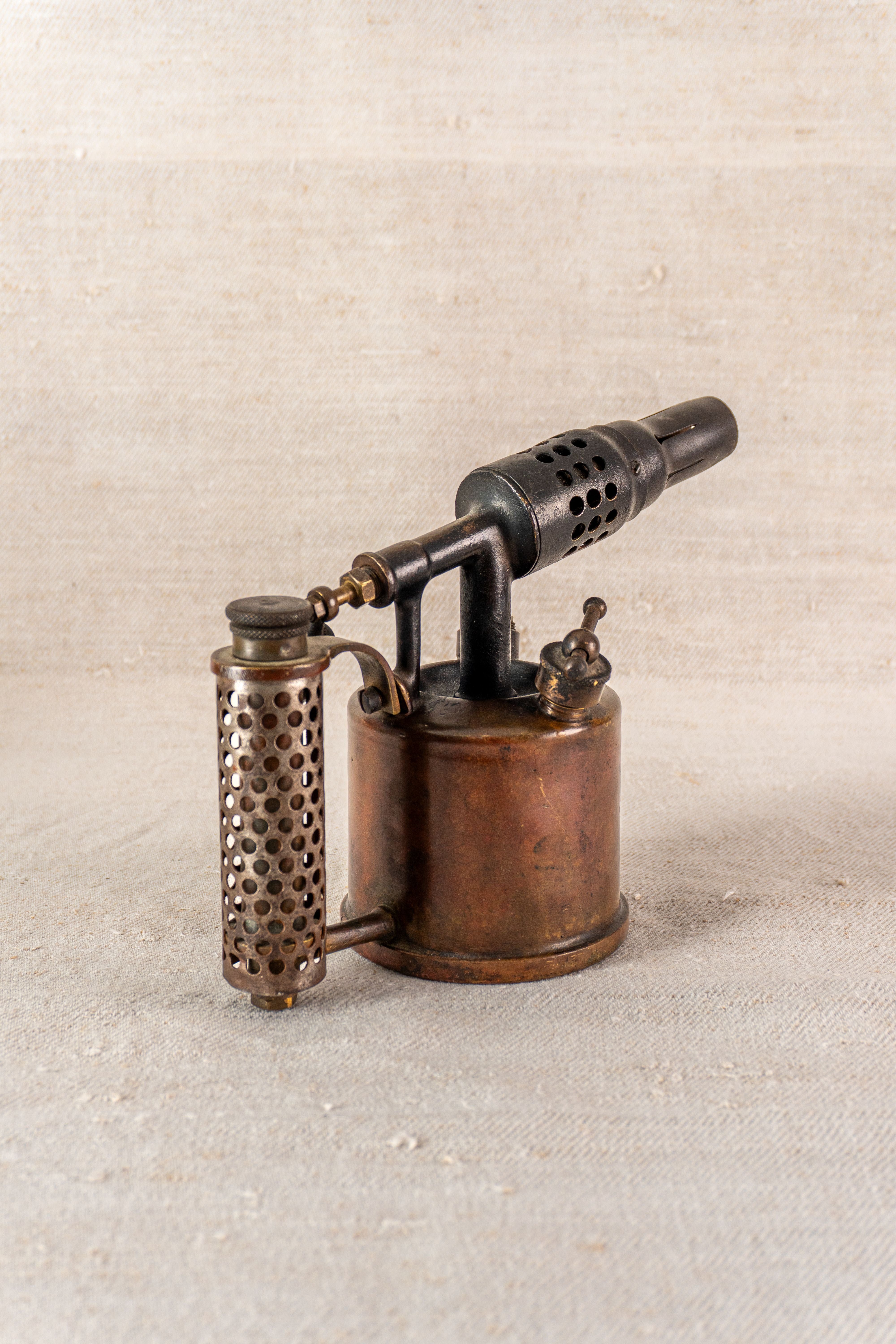 Vintage French Vesta F.J. Paris blowtorch, circa 1920, with perforated handle and round tank on light textured fabric.