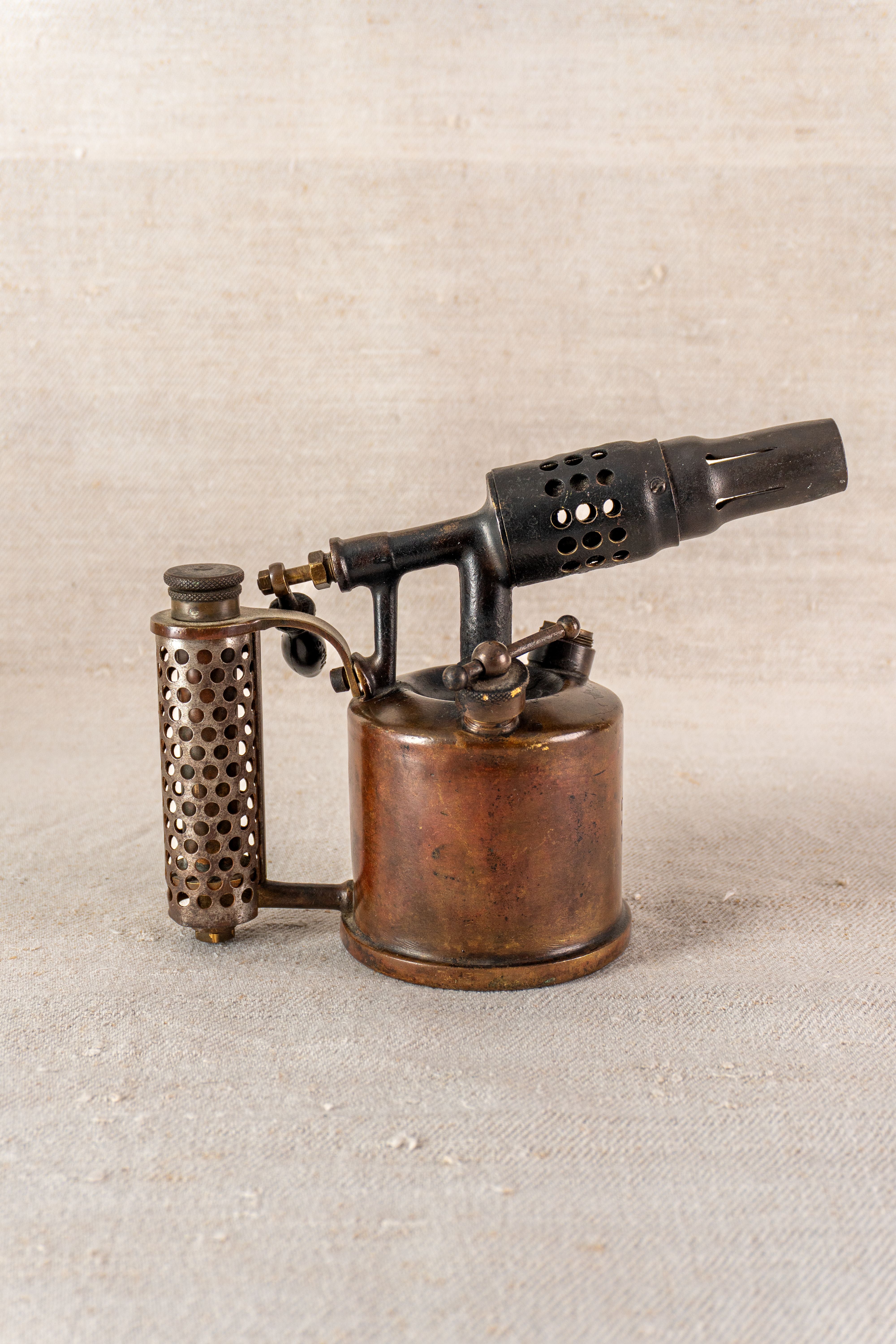 Vintage French blowtorch by Vesta F.J. Paris, circa 1920, cylindrical copper body, black nozzle, on neutral textured surface.