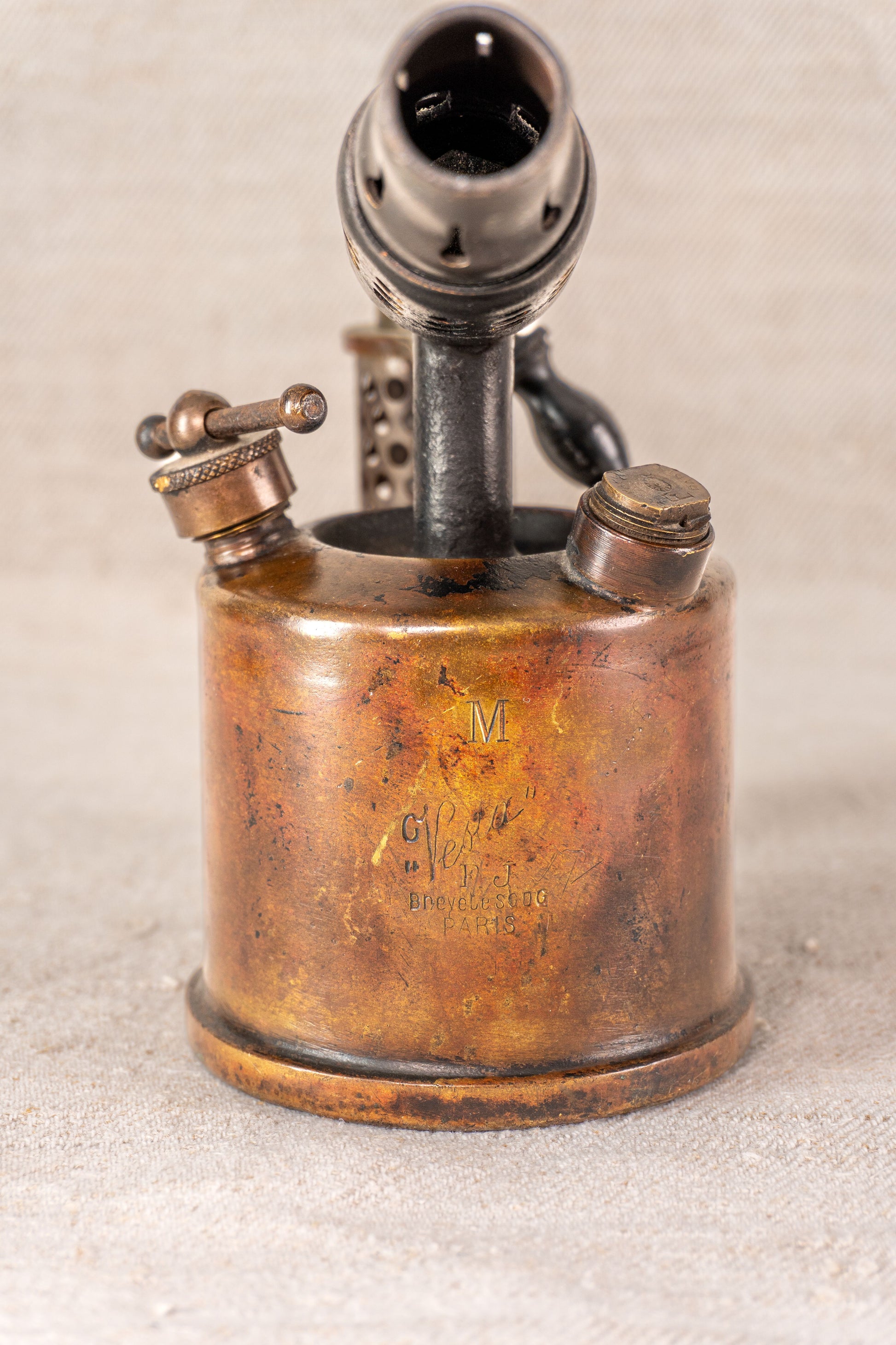 Vintage French Vesta F.J. Paris blowtorch, circa 1920, aged brass with engraved front, on a light textured surface.