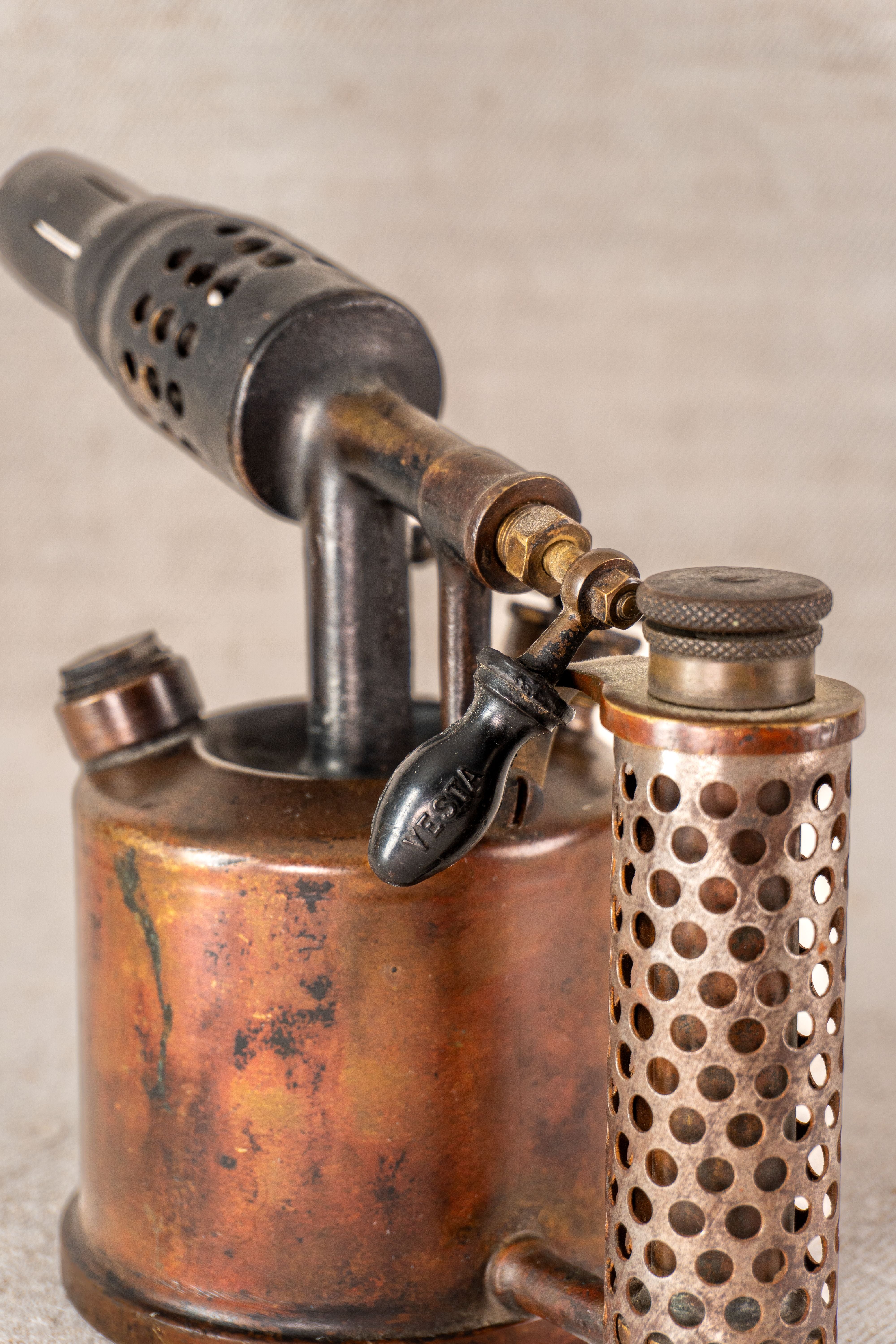 Vintage French Vesta F.J. Paris brass and copper blowtorch with perforated handle, on neutral fabric background.