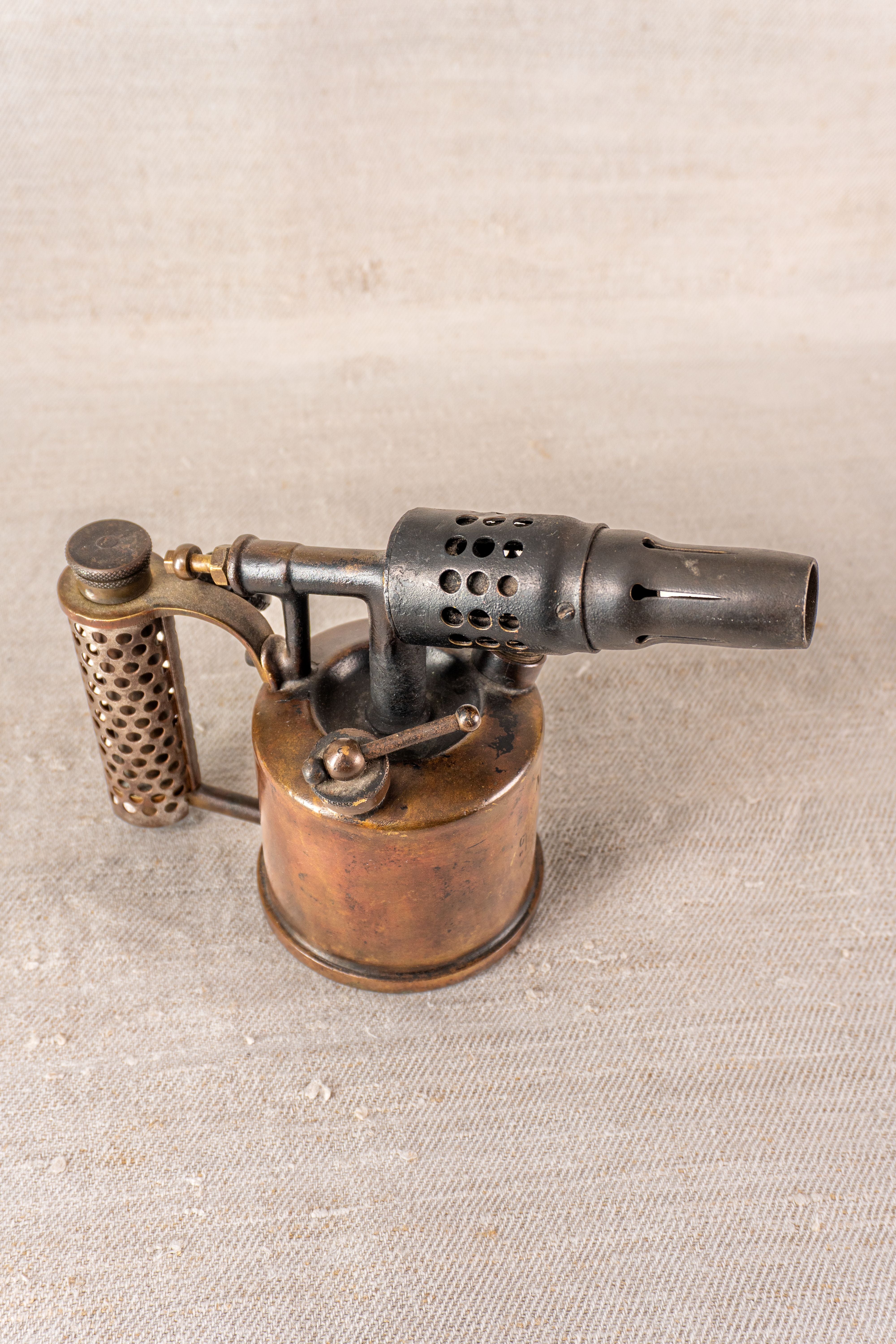 Vintage French Vesta F.J. Paris blowtorch, c.1920, brass tank and vented nozzle on textured beige backdrop.