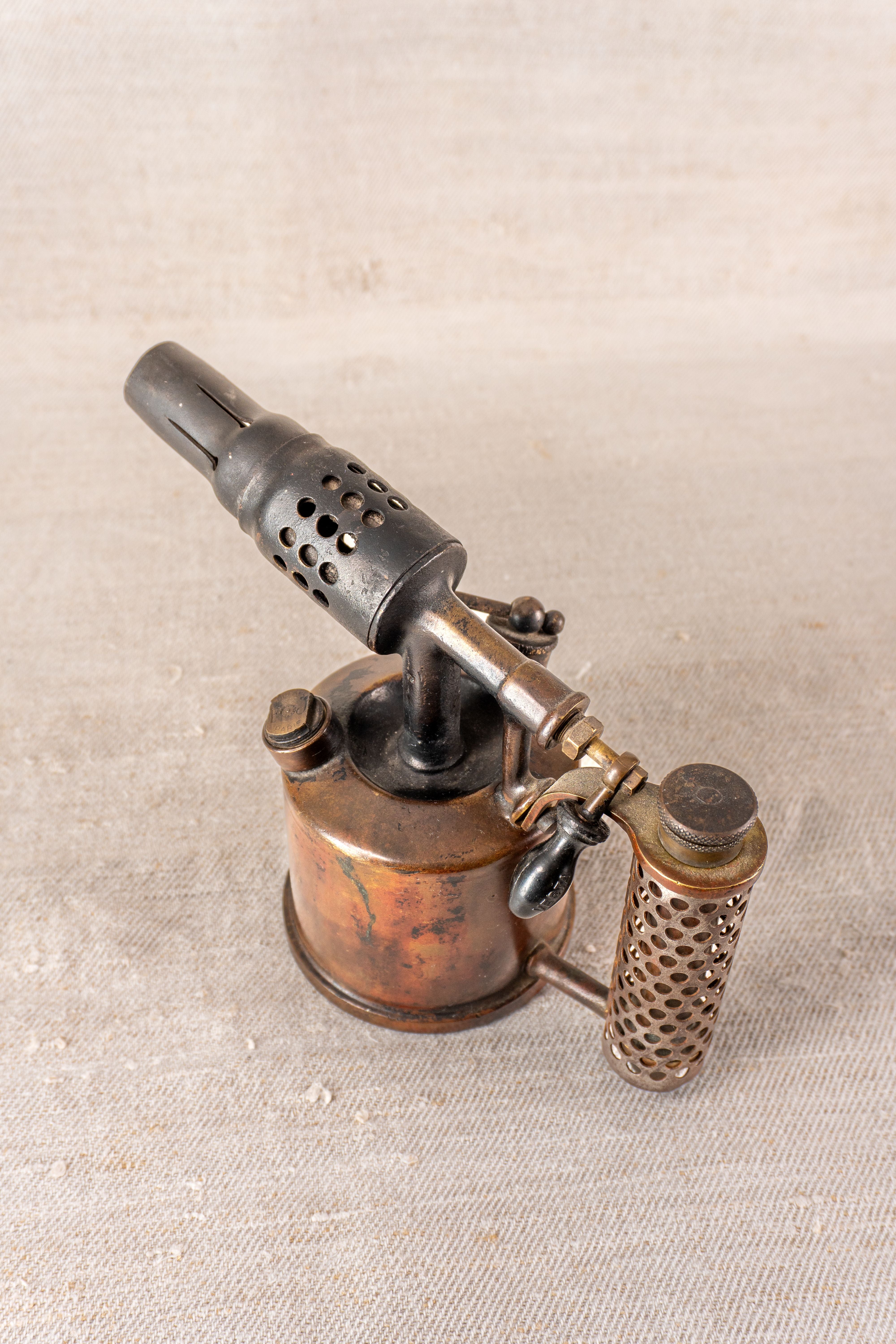 Vintage French blowtorch by Vesta F.J. Paris, circa 1920, brass body with perforated handle, aged patina on light textured surface.