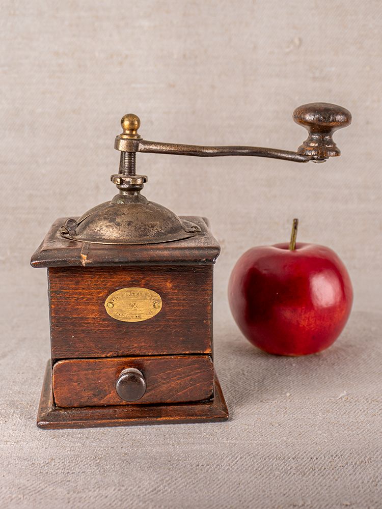 Antique French Peugeot Frères coffee grinder with metal handle beside a red apple on light fabric surface.