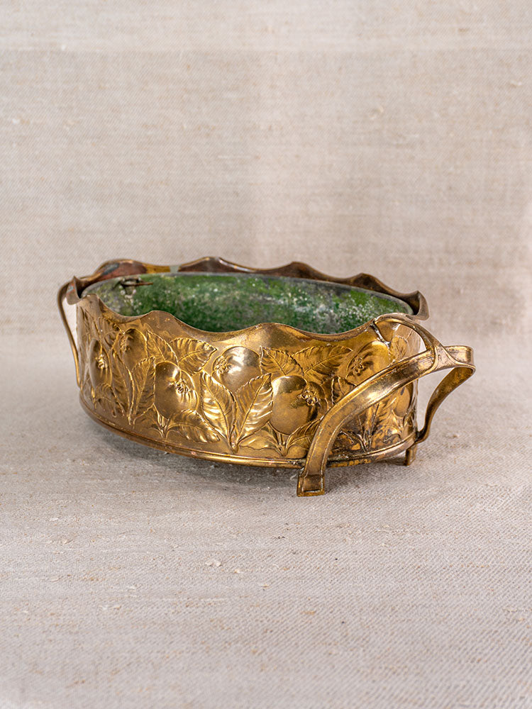 Vintage Swiss brass jardinière with embossed fruit motif, curved handles, green interior on neutral fabric background.