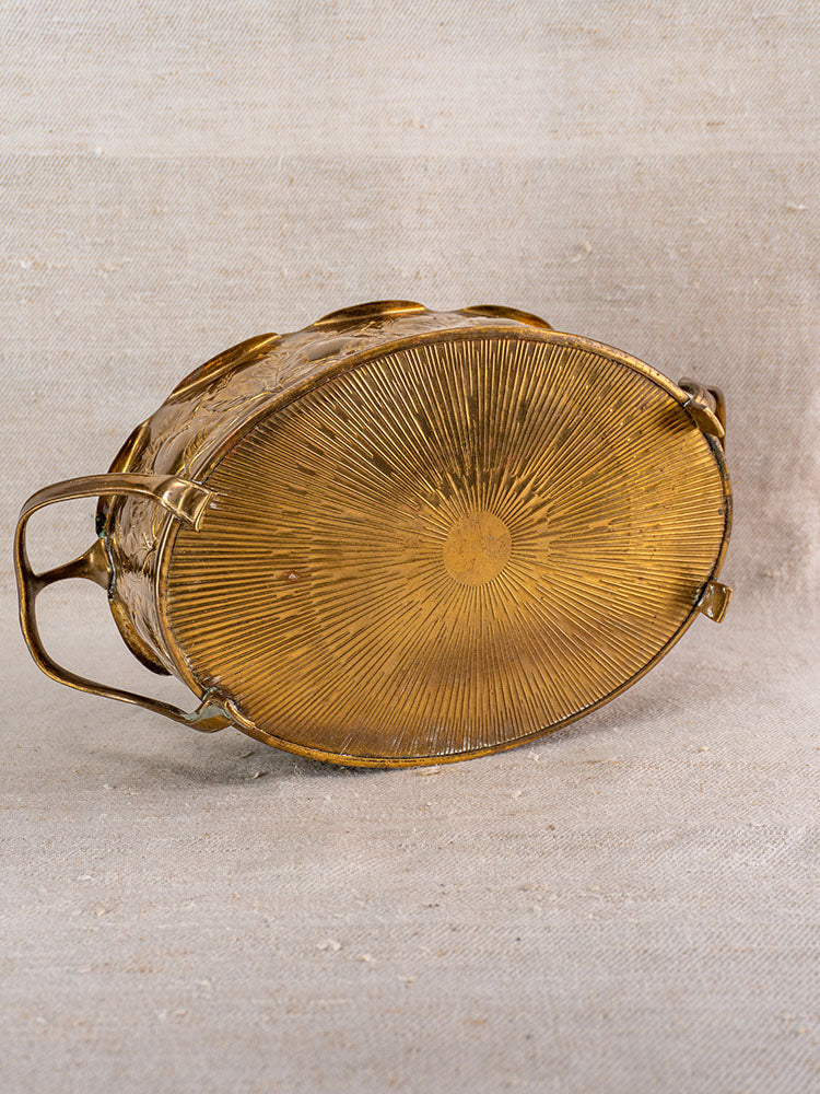 Vintage Swiss brass jardinière upside down, embossed fruit motif and radiating sunburst base on beige fabric.